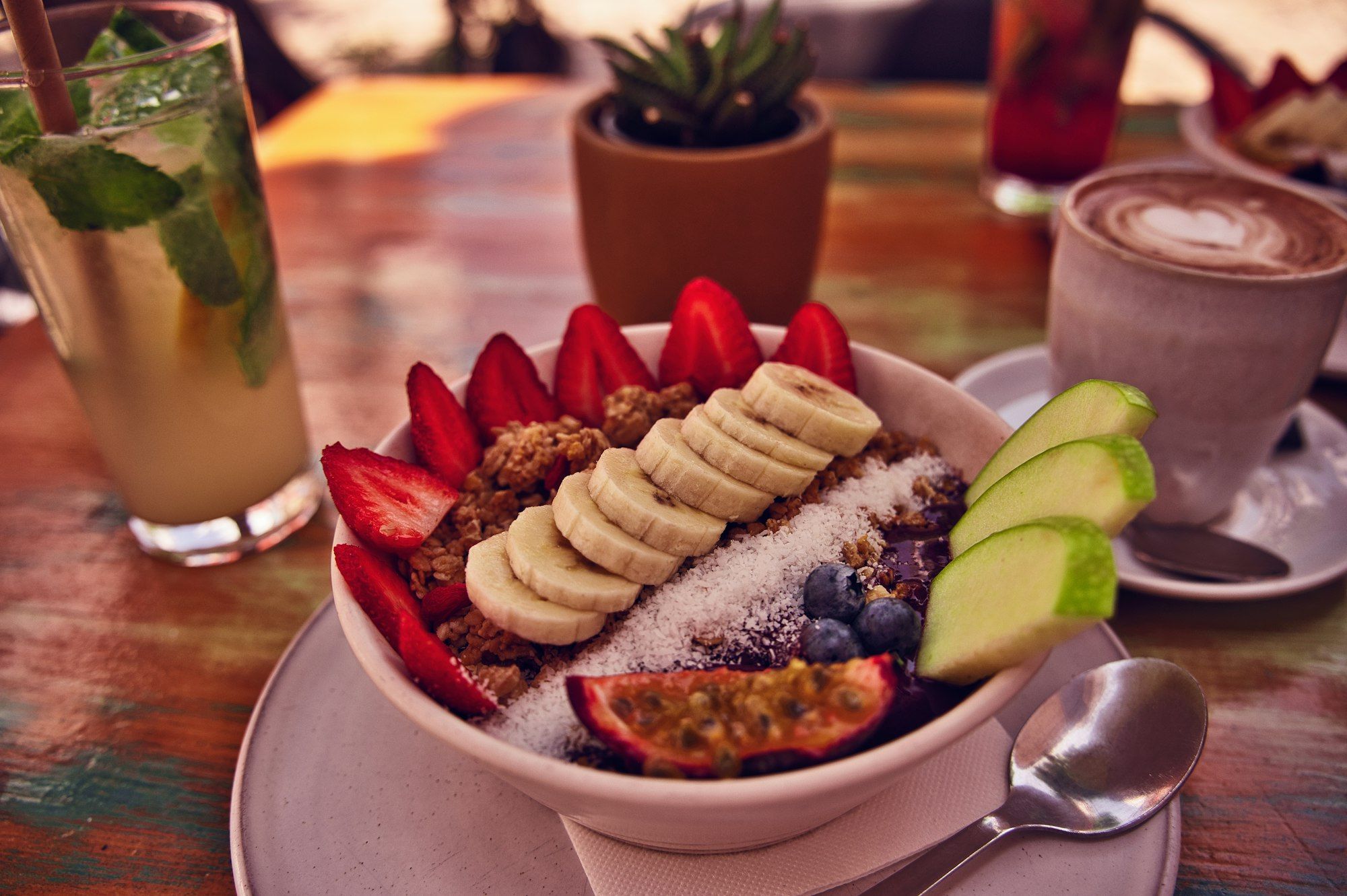Smoothie bowl pitaya dengan pisang, stroberi, granola, dan alpukat, ditemani kopi susu