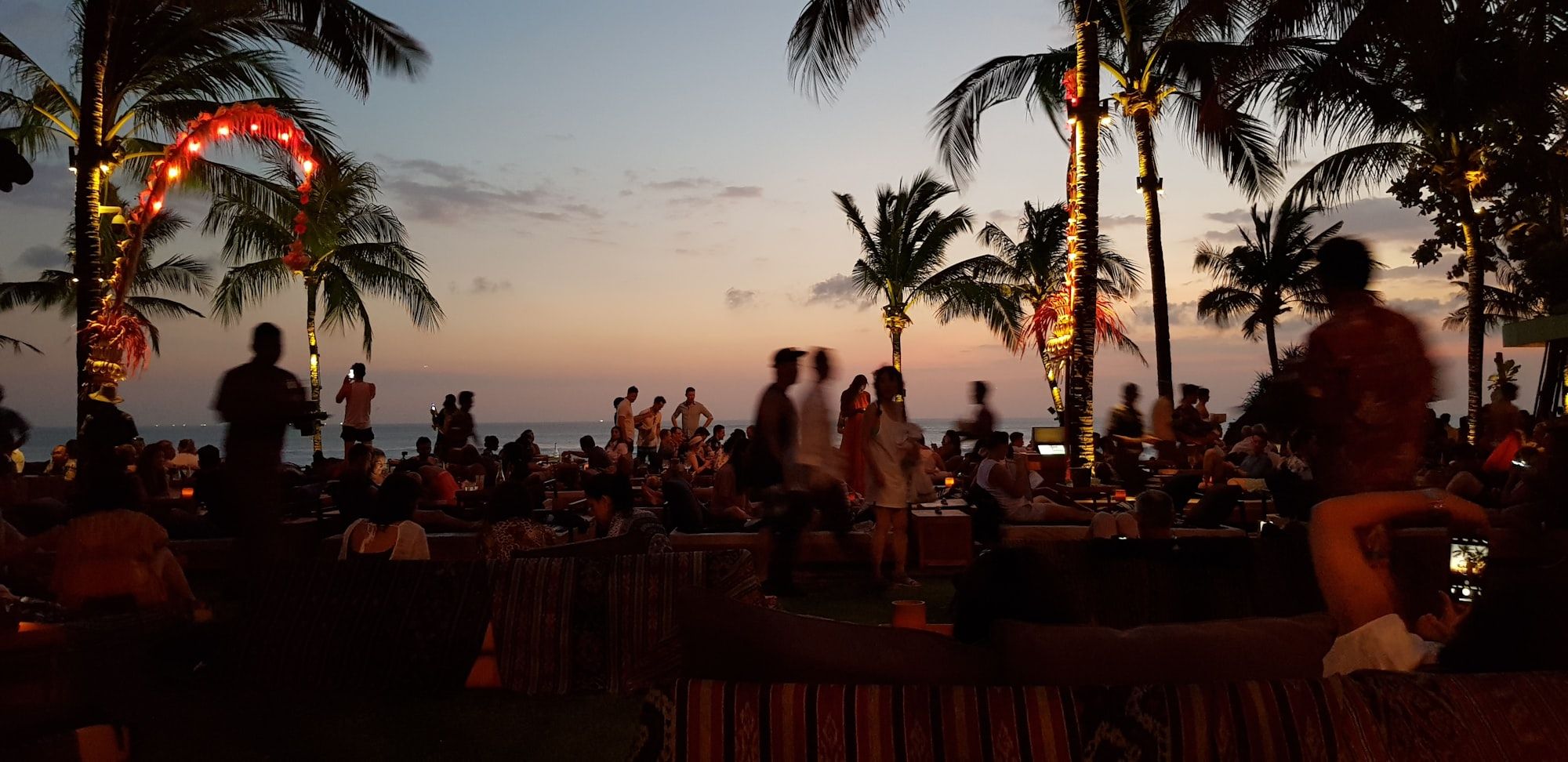 Tamu berkumpul di bar tepi pantai saat senja, lampu tumblr dan pohon kelapa jadi siluet